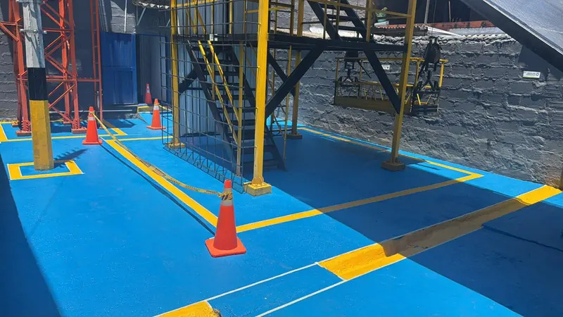 Piso exterior azul con líneas y recuadros amarillos de señalización, conos y escaleras metálicas